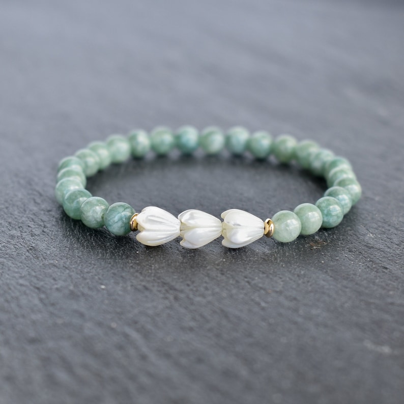 May include: A stretch bracelet featuring round, pale green beads and three white, carved flower-shaped beads at the center. The floral beads are flanked by small, metallic gold-toned spacer beads, creating a balanced and symmetrical design.
