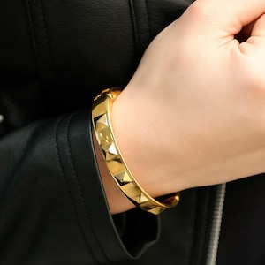 Może przedstawiać: Złota bransoletka o geometrycznym wzorze. Bransoletka posiada szereg ćwieków w kształcie piramid na swojej powierzchni. Biżuteria jest noszona na nadgarstku, a tło stanowi czarna skórzana kurtka.