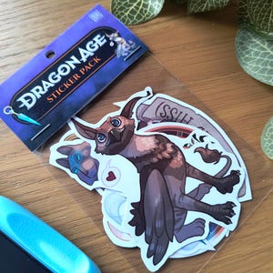 Peut inclure: Un pack de stickers Dragon Age avec une variété d'autocollants colorés illustrant des créatures fantastiques. Les autocollants sont emballés dans un sac en plastique transparent avec une carte d'en-tête bleue et violette qui indique "Dragon Age Sticker Pack".