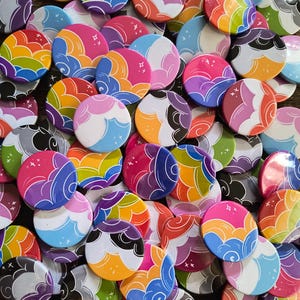 Op de afbeelding: Een verzameling ronde buttons met een herhalend grafisch patroon van gestileerde wolken in diverse kleurencombinaties, waaronder roze, oranje, geel, blauw, paars en zwart, geaccentueerd met kleine witte stervormen.