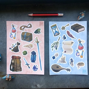 Puede incluir: Dos hojas de pegatinas con temática de fantasía. La hoja de la izquierda es rosa y presenta una mochila, una espada, una linterna, un cofre, una rana, una calavera, un gato y frascos de pociones. La hoja de la derecha es azul y presenta un laúd, un libro, una pluma, una vela, un frasco de poción, un rollo, una llave, un reloj de arena y una rama de lavanda.