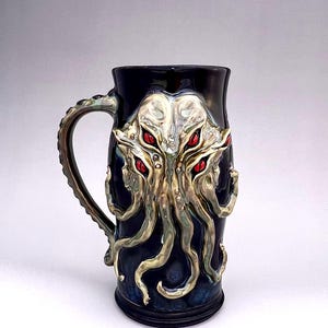 May include: A black ceramic mug with a detailed sculpted design of a multi-eyed, tentacled creature with red eyes. The creature is depicted in a gold and green color scheme.