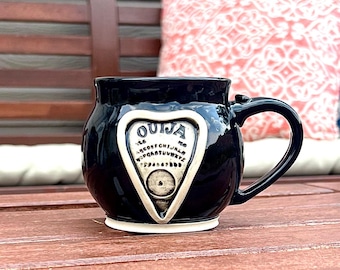 May include: Black ceramic mug with a white heart-shaped design featuring the word "OUIJA" and a classic Ouija board planchette.