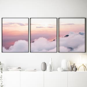 Puede incluir: Tres impresiones enmarcadas de paisajes que representan un amanecer sobre una cordillera, con suaves tonos rosas y morados en el cielo y nubes blancas y esponjosas. La obra de arte se exhibe sobre un mueble blanco con jarrones y una lámpara.