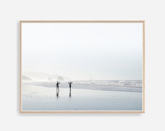 以下が含まれることがあります： 2人の人物がサーフボードを持って砂浜を歩いています。背景には海が広がっています。空は白く霞んでいて、水は薄い青色です。