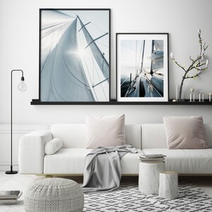 May include: Two framed photographs of a sailboat with white sails against a blue sky. The top photo shows the sail from below, while the bottom photo shows the boat from the deck. The photos are hanging on a white wall above a white couch with gray throw pillows and a gray throw blanket.