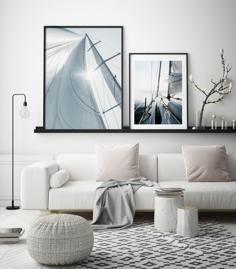 May include: Two framed photographs of a sailboat with white sails against a blue sky. The top photo shows the sail from below, while the bottom photo shows the boat from the deck. The photos are hanging on a white wall above a white couch with gray throw pillows and a gray throw blanket.