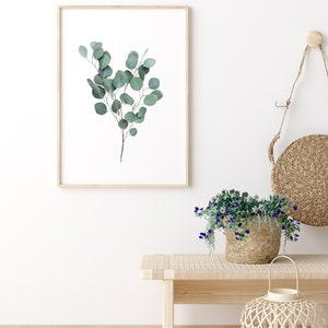 Puede incluir: Un cuadro con una rama de eucalipto con hojas verdes. La obra de arte est&aacute; colgada en una pared blanca sobre un banco de madera. Tambi&eacute;n se ve una cesta de flores moradas y una linterna tejida.