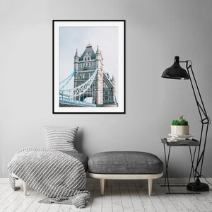 May include: A framed photograph of Tower Bridge in London, England. The bridge is a gray stone structure with blue metal walkways. The photo is in a black frame and is hanging on a gray wall.