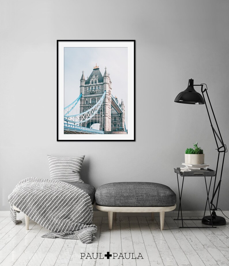 May include: A framed photograph of Tower Bridge in London, England. The bridge is a gray stone structure with blue metal walkways. The photo is in a black frame and is hanging on a gray wall.
