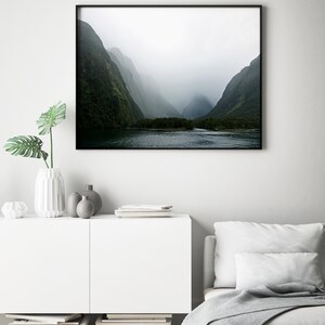 May include: A black and white photograph of a misty mountain valley with a lake in the foreground. The mountains are covered in lush green trees and the sky is a soft gray.