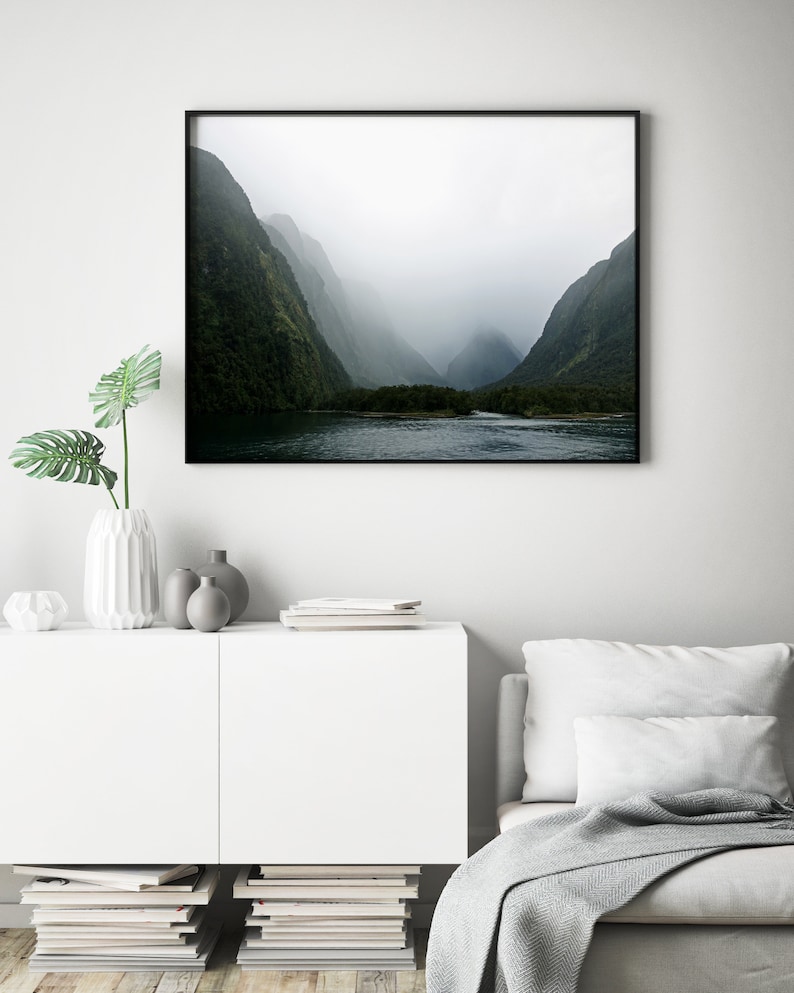May include: A black and white photograph of a misty mountain valley with a lake in the foreground. The mountains are covered in lush green trees and the sky is a soft gray.