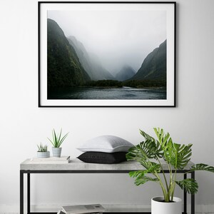 May include: A framed photograph of a misty mountain landscape with a river in the foreground. The photo is hanging on a white wall above a concrete table with a potted plant and two smaller potted plants.