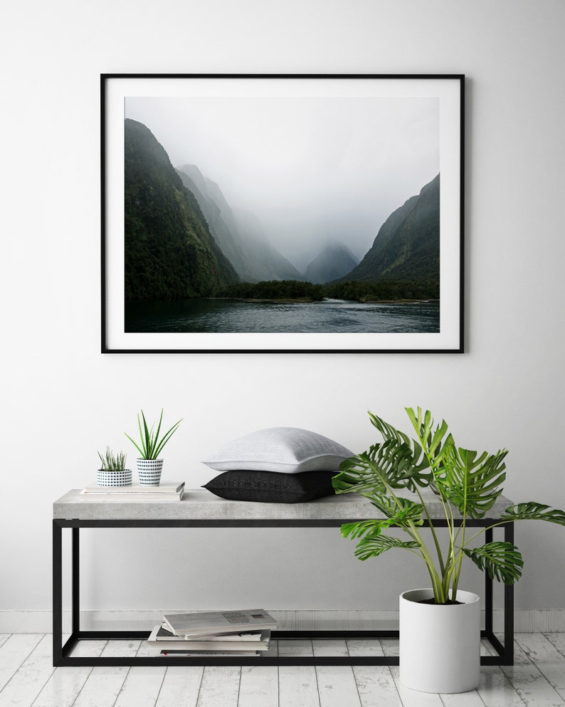 May include: A framed photograph of a misty mountain landscape with a river in the foreground. The photo is hanging on a white wall above a concrete table with a potted plant and two smaller potted plants.