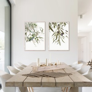 K&ouml;nnte beinhalten: Zwei gerahmte Drucke von Olivenzweigen mit gr&uuml;nen Bl&auml;ttern und Oliven, die von den Zweigen h&auml;ngen. Die Drucke h&auml;ngen an einer wei&szlig;en Wand in einem Esszimmer mit einem Holztisch und wei&szlig;en St&uuml;hlen.