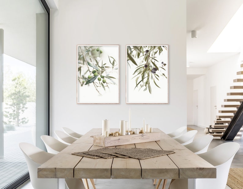 K&ouml;nnte beinhalten: Zwei gerahmte Drucke von Olivenzweigen mit gr&uuml;nen Bl&auml;ttern und Oliven, die von den Zweigen h&auml;ngen. Die Drucke h&auml;ngen an einer wei&szlig;en Wand in einem Esszimmer mit einem Holztisch und wei&szlig;en St&uuml;hlen.