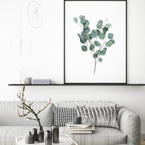 Puede incluir: Impresi&oacute;n enmarcada con una rama de eucalipto con hojas verdes. La obra de arte se exhibe sobre un sof&aacute; gris con cojines decorativos y una mesa de centro blanca. La habitaci&oacute;n est&aacute; decorada en un estilo minimalista.
