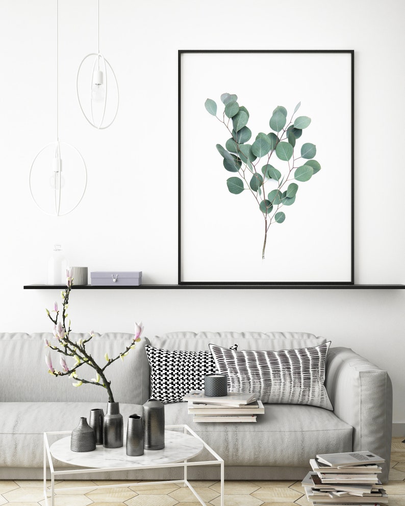 Puede incluir: Impresi&oacute;n enmarcada con una rama de eucalipto con hojas verdes. La obra de arte se exhibe sobre un sof&aacute; gris con cojines decorativos y una mesa de centro blanca. La habitaci&oacute;n est&aacute; decorada en un estilo minimalista.