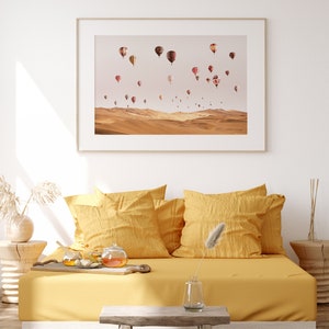 May include: A framed print of a desert landscape with hot air balloons in the sky. The print is hanging on a white wall above a yellow couch with pillows.