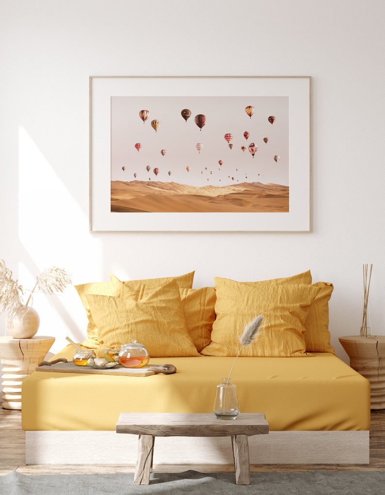 May include: A framed print of a desert landscape with hot air balloons in the sky. The print is hanging on a white wall above a yellow couch with pillows.