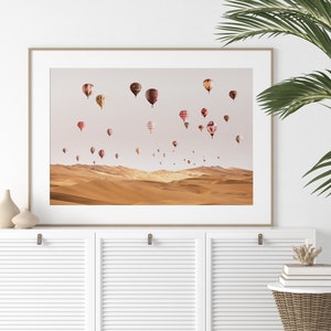 May include: A framed print of a desert landscape with a sky full of colorful hot air balloons. The balloons are various shapes and sizes, and some have patterns on them. The desert is a warm, sandy color.