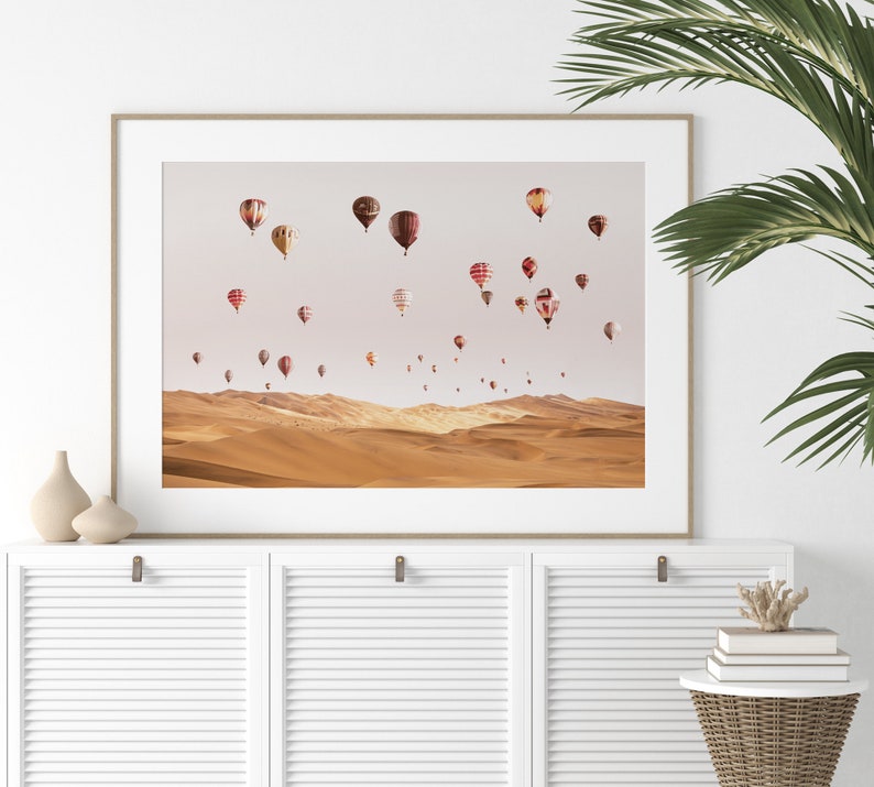 May include: A framed print of a desert landscape with a sky full of colorful hot air balloons. The balloons are various shapes and sizes, and some have patterns on them. The desert is a warm, sandy color.