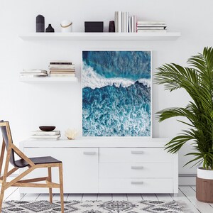 May include: A framed photograph of an aerial view of ocean waves crashing against a rocky shore. The water is a deep blue and the waves are white and foamy. The photo is hanging on a white wall above a white dresser.