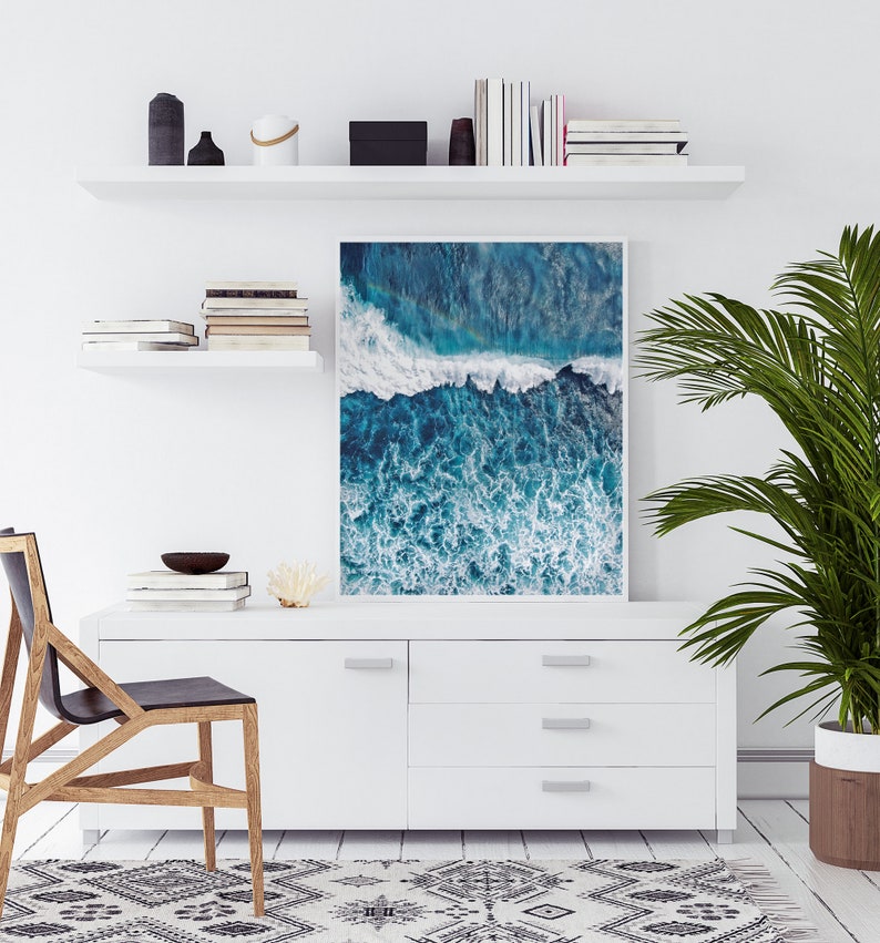 May include: A framed photograph of an aerial view of ocean waves crashing against a rocky shore. The water is a deep blue and the waves are white and foamy. The photo is hanging on a white wall above a white dresser.