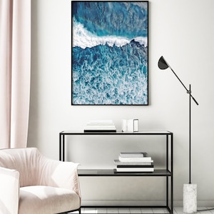 May include: An aerial photograph of a blue ocean with white foamy waves crashing. The image is framed in a black frame and hanging on a white wall.