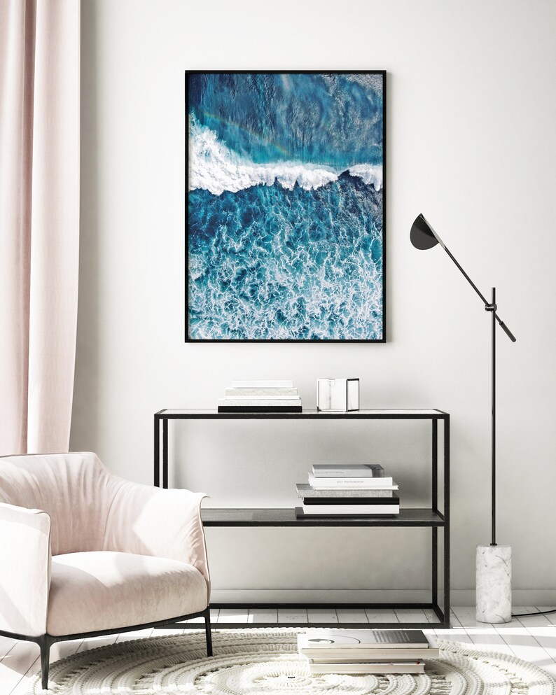 May include: An aerial photograph of a blue ocean with white foamy waves crashing. The image is framed in a black frame and hanging on a white wall.