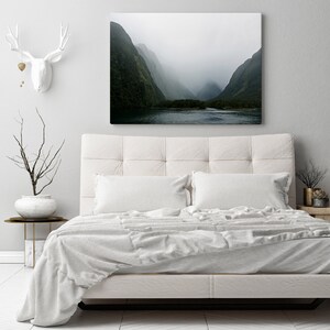 May include: A white bed with a white duvet cover and two white pillows. The bed is in front of a white wall with a framed photograph of a misty mountain landscape.