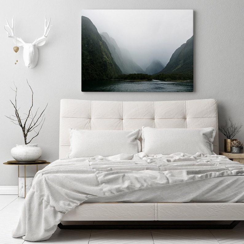 May include: A white bed with a white duvet cover and two white pillows. The bed is in front of a white wall with a framed photograph of a misty mountain landscape.