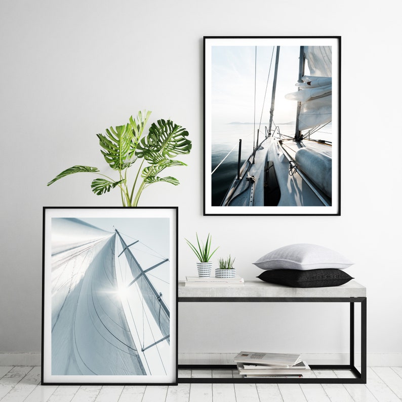 May include: Two framed black and white photographs of sailboats on a white wall. The top photo shows a sailboat with a white sail, viewed from the deck looking forward. The bottom photo shows a close-up of a white sail billowing in the wind.
