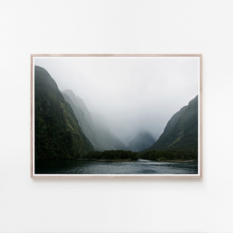 May include: A misty mountain landscape with a body of water in the foreground. The mountains are covered in lush green foliage and the water is a deep blue.
