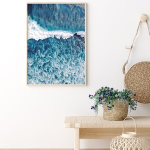 May include: A framed photograph of a blue ocean with white waves crashing. The photo is hanging on a white wall above a wooden bench with a wicker basket of flowers and a wicker lantern.
