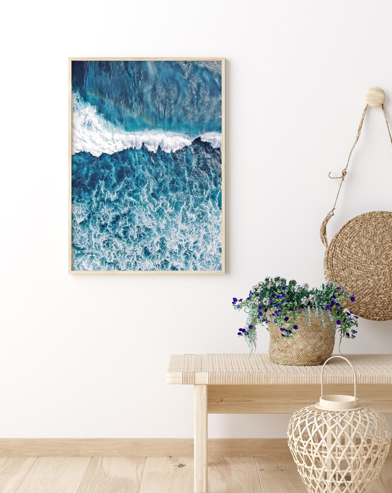 May include: A framed photograph of a blue ocean with white waves crashing. The photo is hanging on a white wall above a wooden bench with a wicker basket of flowers and a wicker lantern.