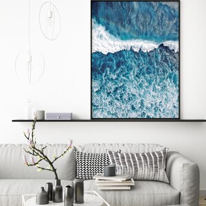 May include: A framed photograph of an aerial view of ocean waves crashing against the shore. The water is a deep blue color with white foam. The photo is hanging on a white wall above a gray couch.