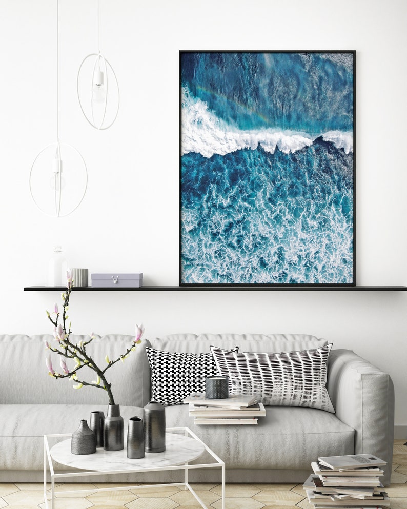 May include: A framed photograph of an aerial view of ocean waves crashing against the shore. The water is a deep blue color with white foam. The photo is hanging on a white wall above a gray couch.