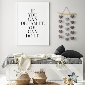 May include: A white framed print with the text "If you can dream it, you can do it." hanging on a white wall above a white daybed with a gray and white striped duvet, a white throw blanket, and two black pillows. There are two woven baskets on the floor in front of the daybed. A wooden branch with a string of gray and purple felt hearts hangs on the wall to the right of the print.