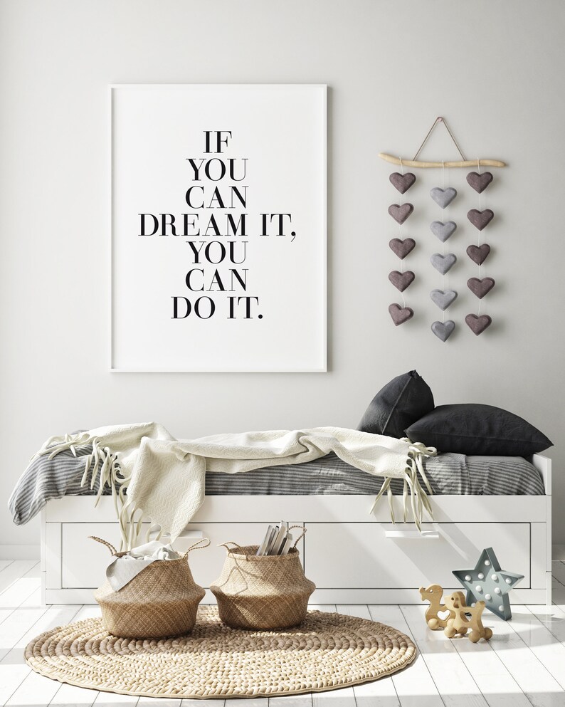 May include: A white framed print with the text "If you can dream it, you can do it." hanging on a white wall above a white daybed with a gray and white striped duvet, a white throw blanket, and two black pillows. There are two woven baskets on the floor in front of the daybed. A wooden branch with a string of gray and purple felt hearts hangs on the wall to the right of the print.