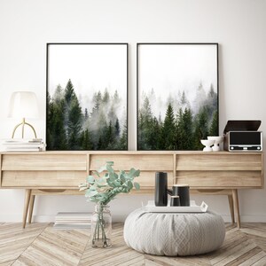 May include: Two black framed prints of a foggy forest scene hang above a light wood console table with two drawers. A vase with green leaves sits on the table in front of the prints. Two black ceramic vases and a white ceramic tray are on the table.