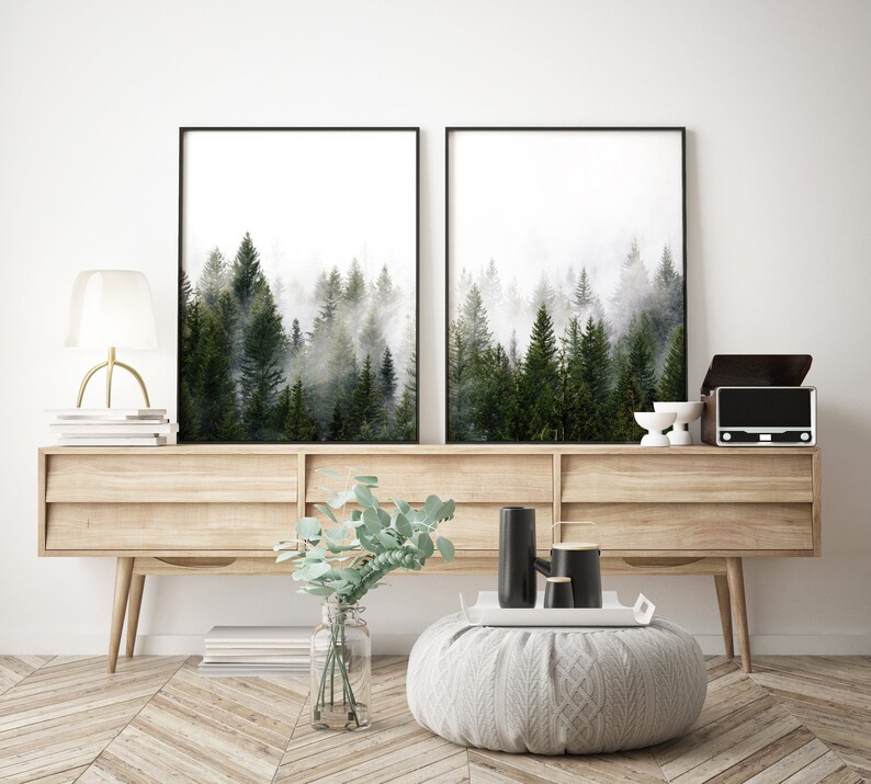 May include: Two black framed prints of a foggy forest scene hang above a light wood console table with two drawers. A vase with green leaves sits on the table in front of the prints. Two black ceramic vases and a white ceramic tray are on the table.