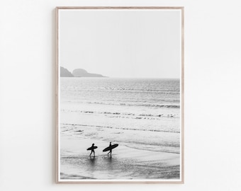 May include: Black and white photograph of two people walking on a beach with surfboards. The people are silhouetted against the horizon, and the sea is in the background.