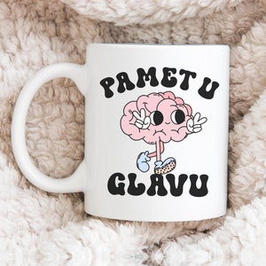 May include: White ceramic mug with a black and white illustration of a brain with eyes and arms. The text "PAMET U GLAVU" is printed on the mug.