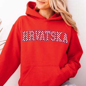 May include: A red hooded sweatshirt with the word "HRVATSKA" in a checkered pattern across the chest. The sweatshirt has a front pocket and a drawstring hood. The person is wearing blue jeans.