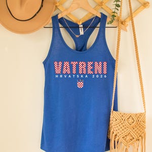 May include: A blue tank top with the word "VATRENI" in red and white checkered letters. Below the word is "HRVATSKA 2026" and a Croatian coat of arms. A tan hat and a woven purse hang nearby.