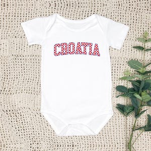 May include: White baby bodysuit with the word "CROATIA" in red and white checkered letters. The bodysuit has short sleeves and a round neckline. The text is in a bold, playful font, and the overall design is simple and clean.
