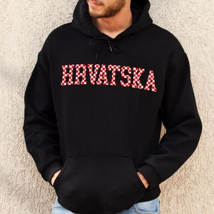 May include: Black hooded sweatshirt with the word "HRVATSKA" in red and white checkered letters across the chest. The sweatshirt has a front pocket and a drawstring hood.