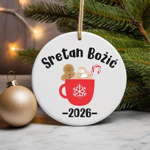 May include: A white ceramic Christmas ornament with the text "Sretan Božić" in black script. A red mug with a snowflake design contains a gingerbread man, marshmallows, and a candy cane. The year "2026" is below the mug. A gold ornament is to the left.