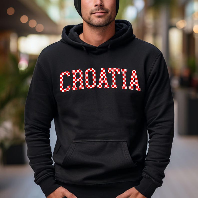 May include: Black hooded sweatshirt with the word "CROATIA" in a red and white checkered pattern across the chest. The sweatshirt has a front pocket and a drawstring hood. The person in the image is wearing a black beanie.